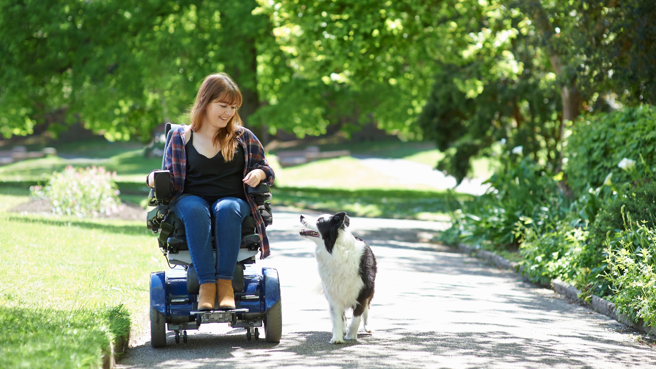 A woman using a power wheelchair shares a smile with a black and white border collie as they travel down a paved path, through lush green grass and a canopy of trees.