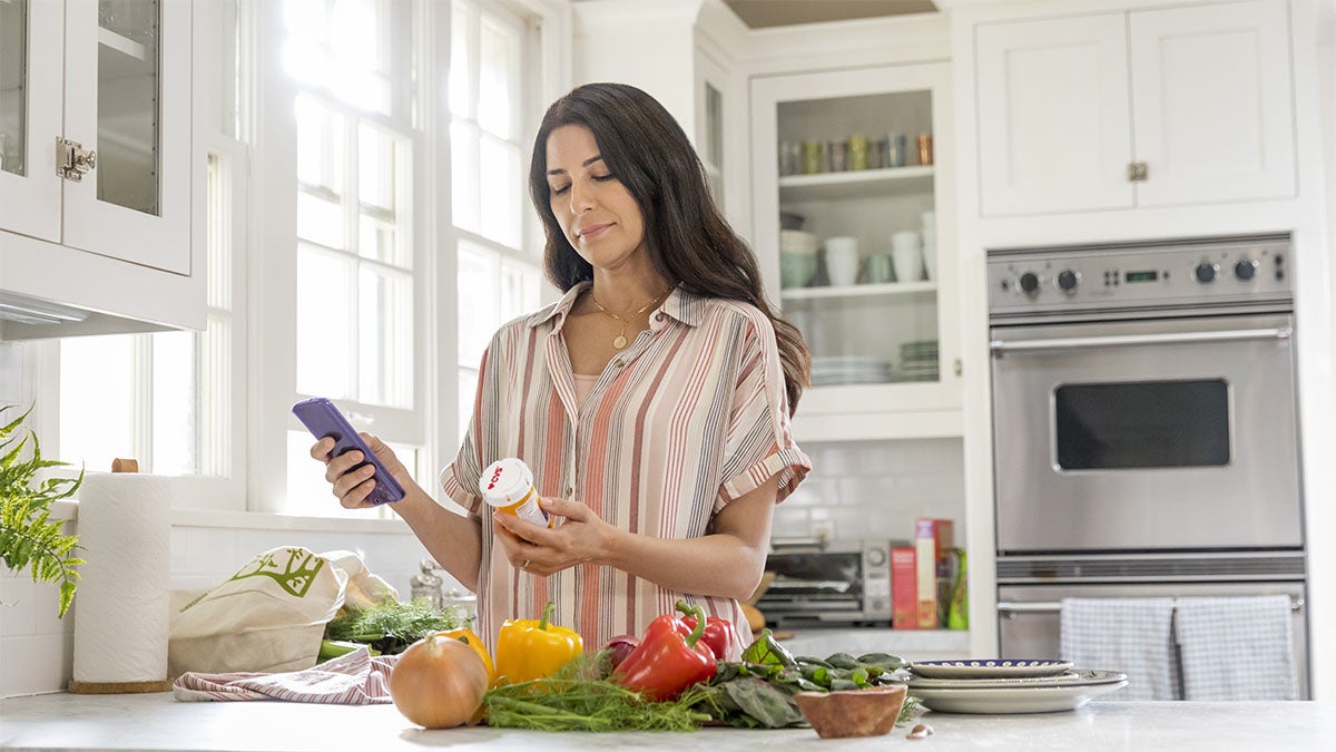 A woman reading a prescription bottle in her kitchen while holding her phone